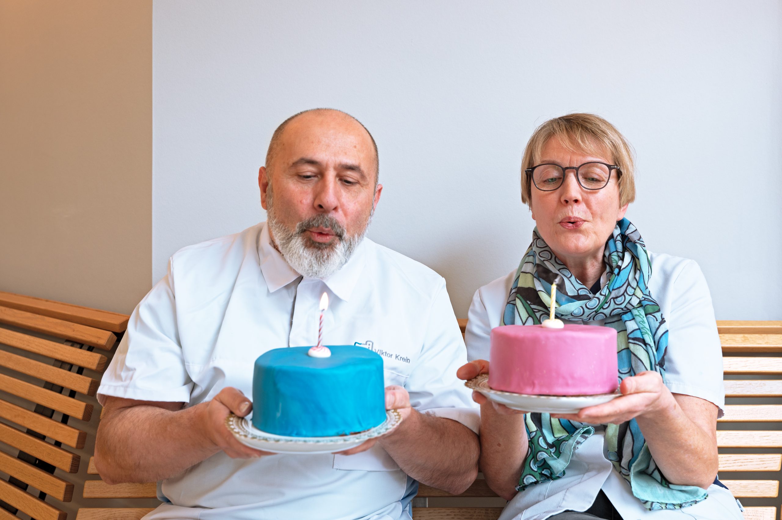 Zwei Mitarbeiter der Praxis halten jeweils einen Kuchen in verschiedenen Fraben in den Händen und pusten die Kerze die Kerze aus.