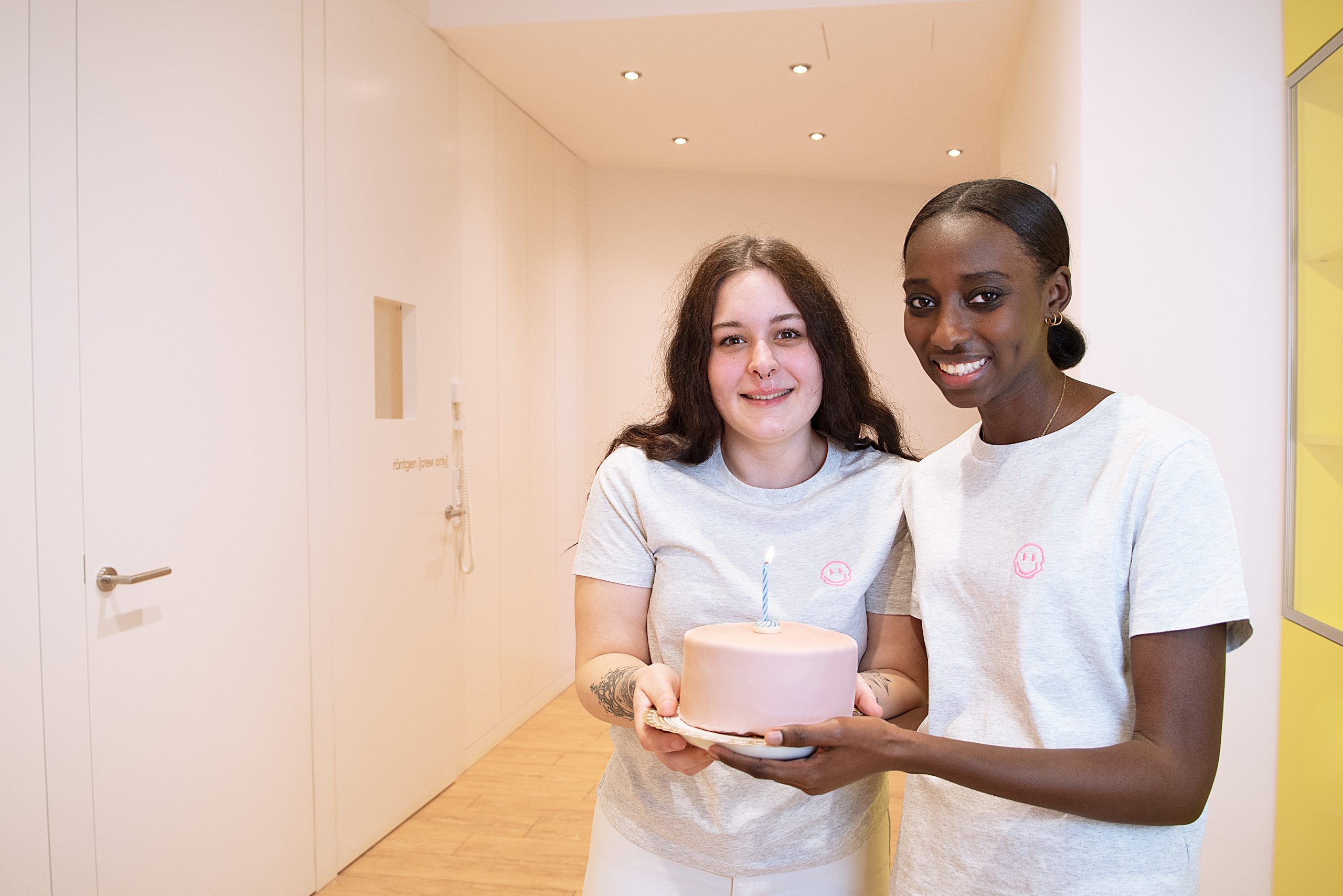 Zwei Mitarbeiterinnen der Praxis halten einen rosanen Kuchen in den Händen und lächeln.